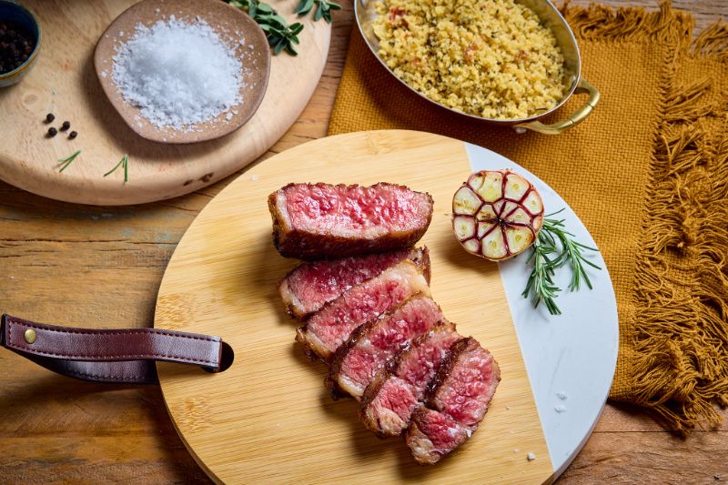 Close-up de um bife de Wagyu perfeitamente cozido em uma tábua de cortar de madeira, decorado com ramos de alecrim, sal grosso e pimenta-do-reino, com um pequeno prato de molho ao lado.