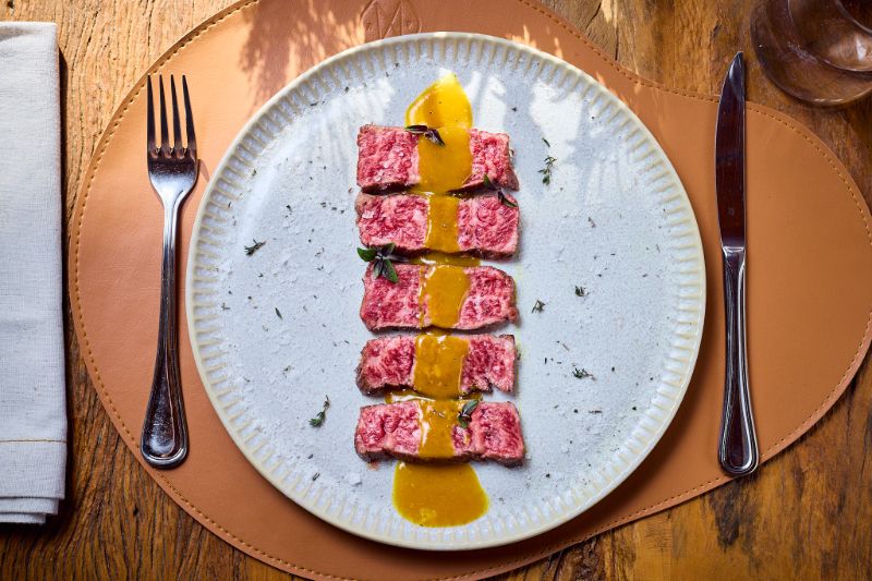 Carne Wagyu suculenta em prato grande central mostrando marmoreio, acompanhada de faca de chef, garfo e potes de temperos sobre mesa de madeira clara.